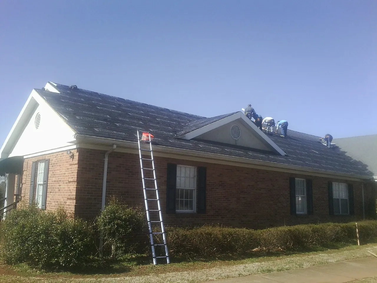 Crew tearing shingles off a brick ranch roof with a ladder set — Metal Roof Repair work by our crew in Iva