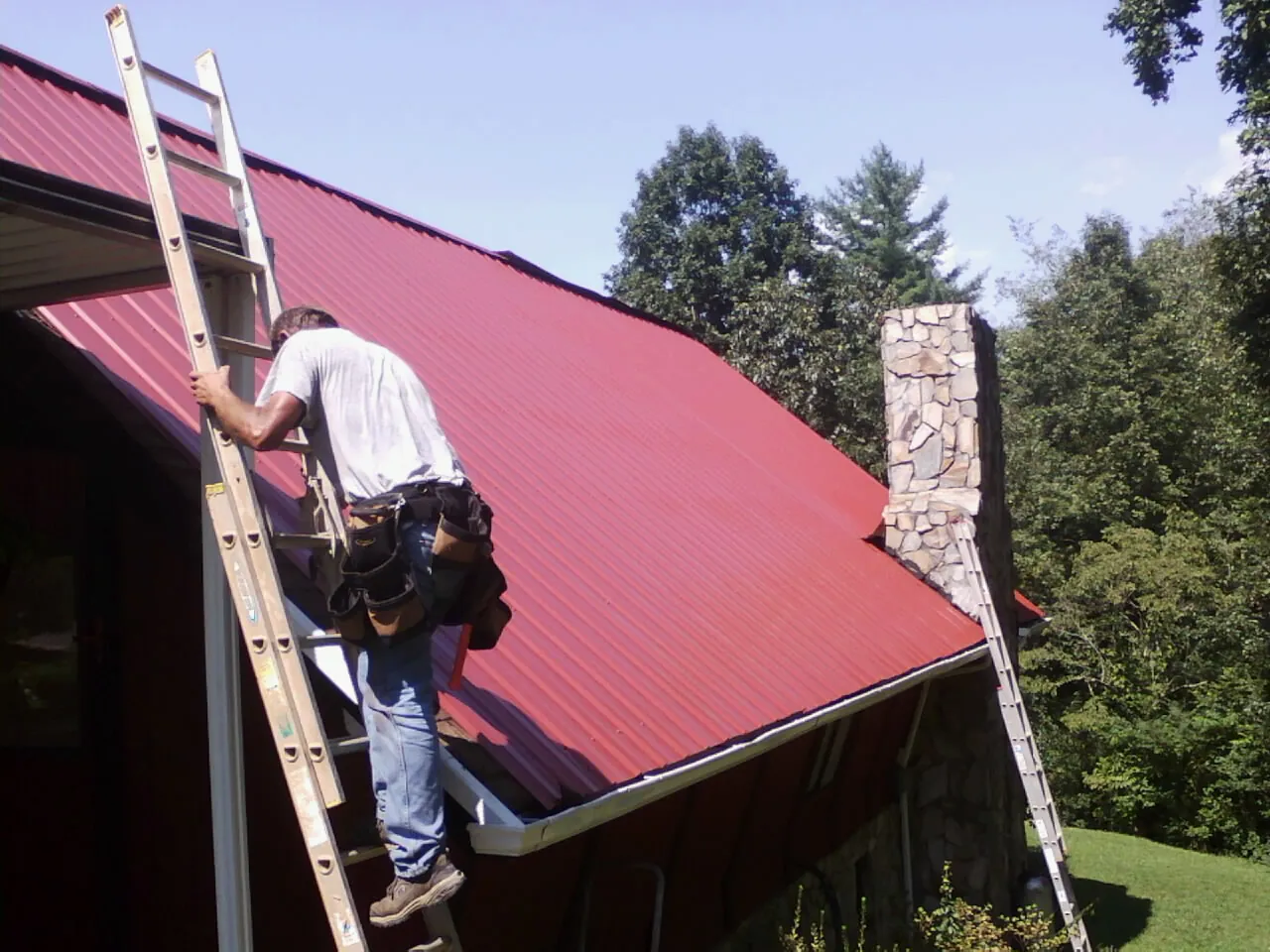 Roofer on a ladder beside a red metal roof and stone chimney — Soffit & Fascia Repair by Roofing Brothers in Iva