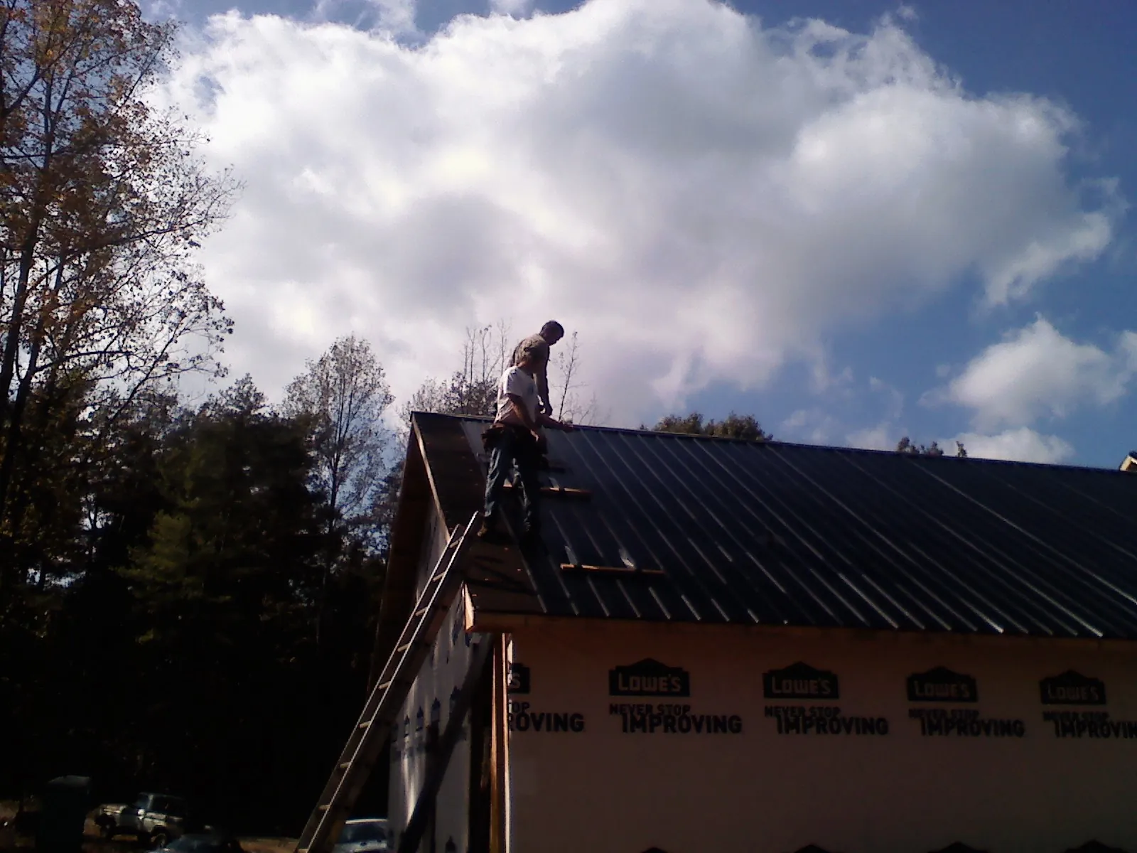 Roofer fastening dark metal panels high against a cloudy sky, from a Roof Inspection job in Iva